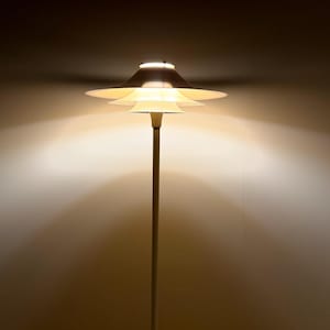 K&ouml;nnte beinhalten: Eine moderne Stehlampe mit einem mehrstufigen, scheibenf&ouml;rmigen Schirm. Die Lampe hat einen schlanken, vertikalen Pfosten und strahlt ein warmes, diffuses Licht aus. Der Schirm besteht aus mehreren &uuml;berlappenden, kreisf&ouml;rmigen Elementen, wodurch ein geschichteter Effekt entsteht.