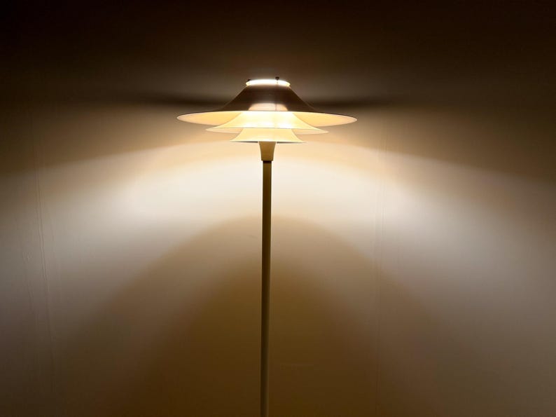 K&ouml;nnte beinhalten: Eine moderne Stehlampe mit einem mehrstufigen, scheibenf&ouml;rmigen Schirm. Die Lampe hat einen schlanken, vertikalen Pfosten und strahlt ein warmes, diffuses Licht aus. Der Schirm besteht aus mehreren &uuml;berlappenden, kreisf&ouml;rmigen Elementen, wodurch ein geschichteter Effekt entsteht.