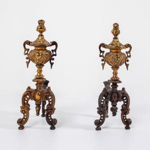 Puede incluir: Un par de morillos de chimenea de latón antiguo a juego con volutas intrincadas y ornamentadas, remates decorativos en forma de urna y bases de trípode resistentes. Cada morillo de metal muestra un acabado dorado desgastado con relieves detallados.