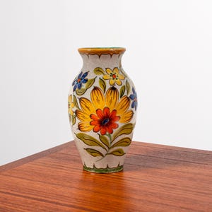 Może przedstawiać: Ceramiczny wazon z kwiatowym wzorem, z żółtymi, czerwonymi i niebieskimi kwiatami na beżowym tle. Wazon, o wysokości około 15 cm, ma zieloną obwódkę i stoi na drewnianej powierzchni.