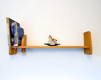 May include: A wooden shelf with a rocking horse toy on it. The shelf is mounted to a white wall. The shelf has a book titled "Vermeer" on it.
