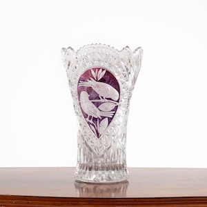 Puede incluir: Un jarrón de cristal tallado transparente con borde festoneado y cuerpo con patrón de diamantes. Un panel ovalado central muestra un fondo morado intenso con un diseño blanco grabado de dos pájaros posados en una rama entre hojas y flores.