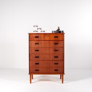 May include: A tall, mid-century modern wooden dresser with six drawers. The dresser is made of reddish-brown wood and has a rectangular shape with a lighter-colored top drawer. Small decorative items sit on top.