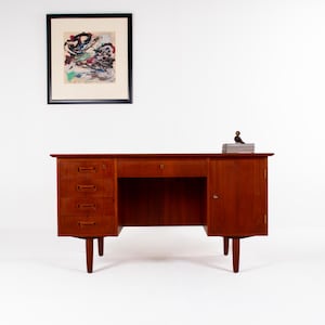 May include: A mid-century modern wooden desk with a warm, reddish-brown finish. The desk features three drawers on the left, a central drawer, and a cabinet on the right. A framed abstract artwork hangs above the desk. A small decorative object sits on top of the desk.