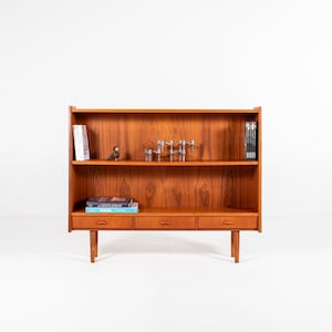 May include: A mid-century modern wooden bookcase with three drawers and two shelves. The bookcase is made of a warm-toned wood and features a minimalist design. The shelves hold books and decorative items.