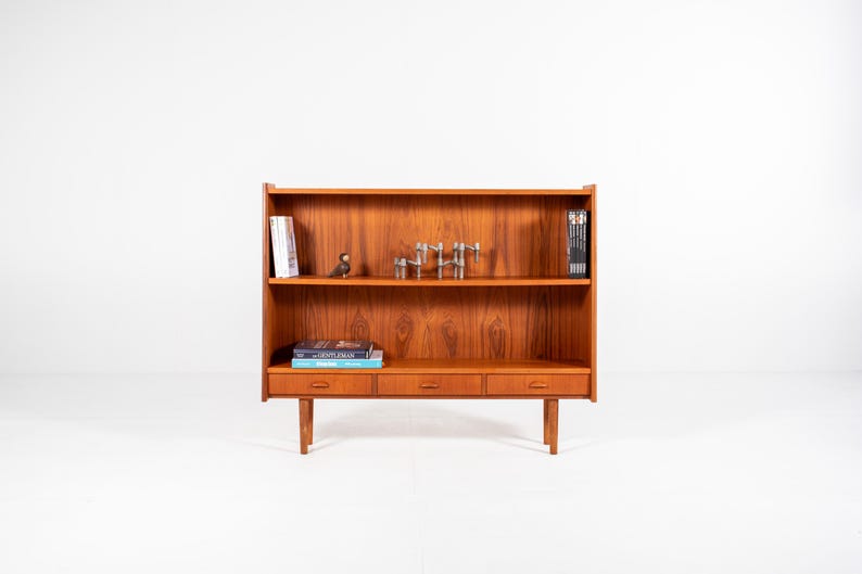 May include: A mid-century modern wooden bookcase with three drawers and two shelves. The bookcase is made of a warm-toned wood and features a minimalist design. The shelves hold books and decorative items.