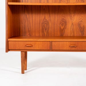 May include: A mid-century modern wooden bookcase with a warm, reddish-brown finish. The bookcase features two shelves, a lower storage area with drawers, and a minimalist design. The wood grain is prominently displayed.