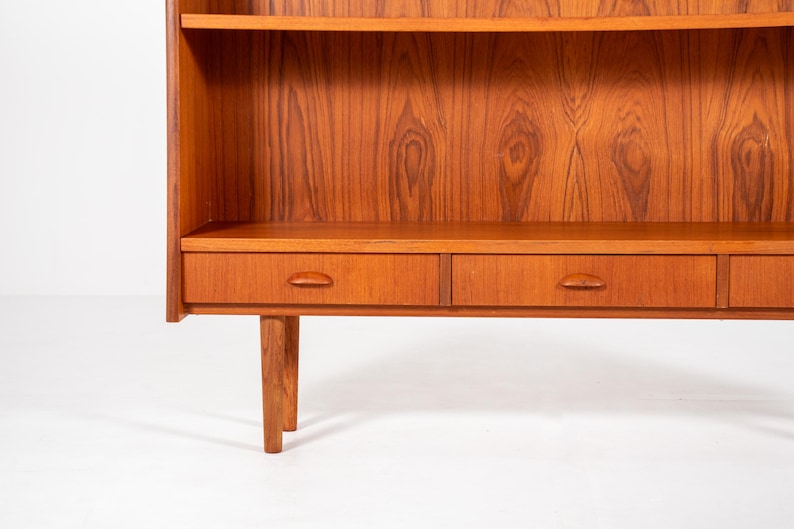 May include: A mid-century modern wooden bookcase with a warm, reddish-brown finish. The bookcase features two shelves, a lower storage area with drawers, and a minimalist design. The wood grain is prominently displayed.