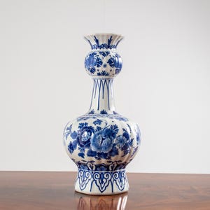 May include: A white ceramic vase with a blue floral pattern, featuring a bulbous base, a slender neck with a rounded mid-section, and a flared rim. The surface is decorated with intricate blue painted flowers and leaves, with a scalloped base design.