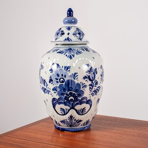 Op de afbeelding: Een witte keramische gemberpot met een afneembaar deksel, voorzien van ingewikkelde blauwe bloem- en bladpatronen geschilderd over het ronde lichaam en deksel. De pot heeft een bolvorm met een taps toelopende hals en een knopvormige bovenkant.