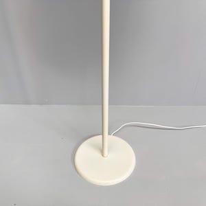 K&ouml;nnte beinhalten: Eine cremefarbene Stehlampe mit rundem Fu&szlig; und geradem Pfosten. Die Lampe hat ein schlichtes, minimalistisches Design und ein wei&szlig;es Stromkabel. Die Lampe steht auf einer grauen Oberfl&auml;che.