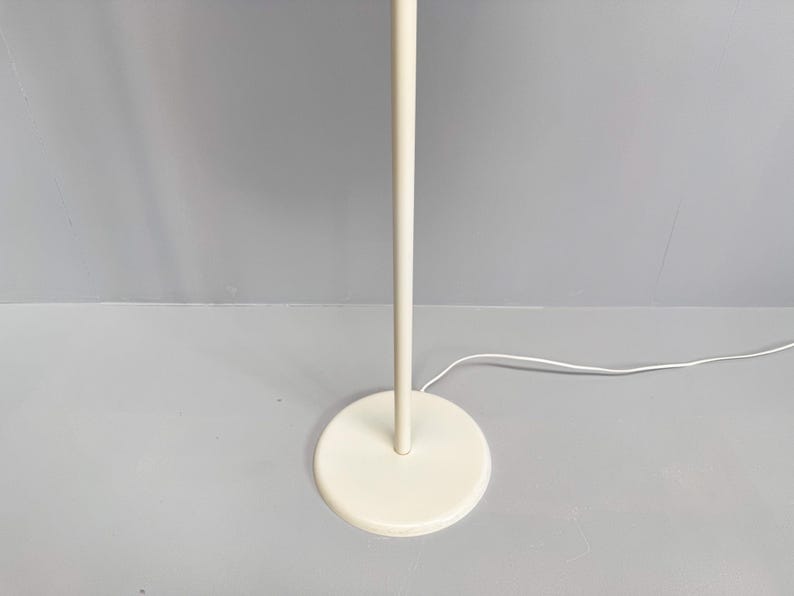 K&ouml;nnte beinhalten: Eine cremefarbene Stehlampe mit rundem Fu&szlig; und geradem Pfosten. Die Lampe hat ein schlichtes, minimalistisches Design und ein wei&szlig;es Stromkabel. Die Lampe steht auf einer grauen Oberfl&auml;che.