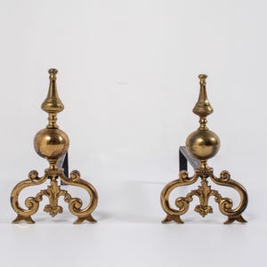 May include: A pair of brass fireplace andirons featuring a central spherical finial topped with a pointed spire, supported by a decorative scrolled base with ornate metalwork details and a dark iron horizontal bar extending towards the rear.