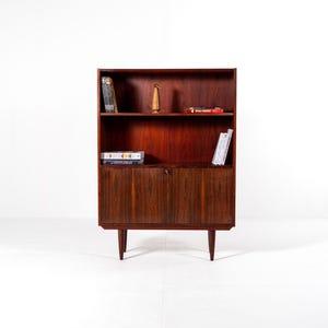 Op de afbeelding: Een houten boekenkast in mid-century modern stijl met een donkerbruine afwerking. De boekenkast heeft planken en een onderkast met een deur. Boeken en decoratieve items worden op de planken tentoongesteld.
