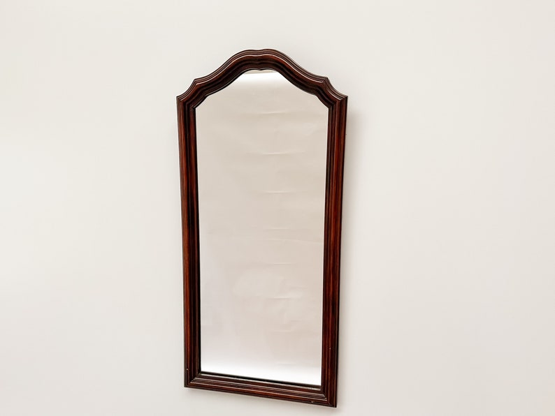 May include: A tall, rectangular mirror with a dark wood frame. The frame has a decorative, arched top with a scalloped edge.