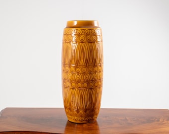 May include: A tall, cylindrical ceramic vase with a glossy amber-brown glaze. The exterior surface features a repeating embossed pattern consisting of horizontal bands of circles and vertical geometric shapes that wrap around the entire body.