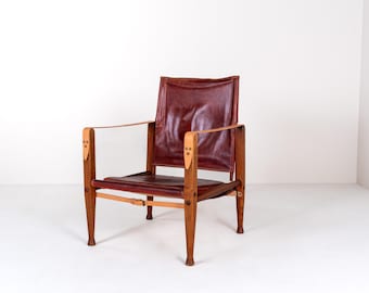 May include: A vintage armchair with a burgundy leather seat and backrest. The chair has a wooden frame with light brown armrests and legs. The design features leather straps and rivets, showcasing a mid-century modern style.