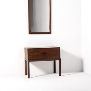 May include: A dark brown wooden nightstand with two drawers and a circular handle. Above it, a rectangular mirror with a matching dark brown frame hangs on the white wall. The furniture has a mid-century modern design.