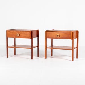 Könnte beinhalten: Zwei passende Nachttische aus Holz mit warmbraunem Finish. Jeder Nachttisch hat eine einzelne Schublade mit einem goldfarbenen Knopf und ein unteres Regal. Die Tische werden von vier Beinen getragen.