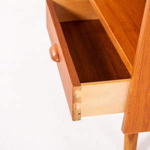 May include: Close-up of a mid-century modern wooden nightstand with an open drawer. The nightstand is made of light brown wood and features a curved drawer pull. The drawer is open, revealing the interior construction.