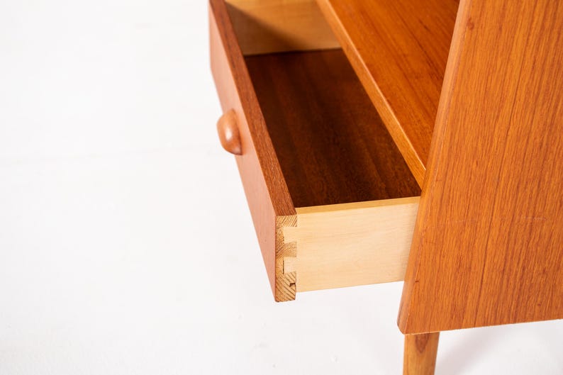 May include: Close-up of a mid-century modern wooden nightstand with an open drawer. The nightstand is made of light brown wood and features a curved drawer pull. The drawer is open, revealing the interior construction.