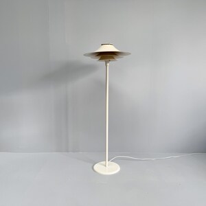 K&ouml;nnte beinhalten: Eine cremefarbene Stehlampe mit einem mehrstufigen Schirmdesign. Die Lampe hat einen schlanken, vertikalen Pfosten und einen kreisf&ouml;rmigen Sockel. Der Schirm besteht aus mehreren gestapelten, scheibenf&ouml;rmigen Elementen. Die Lampe ist an eine Steckdose angeschlossen.