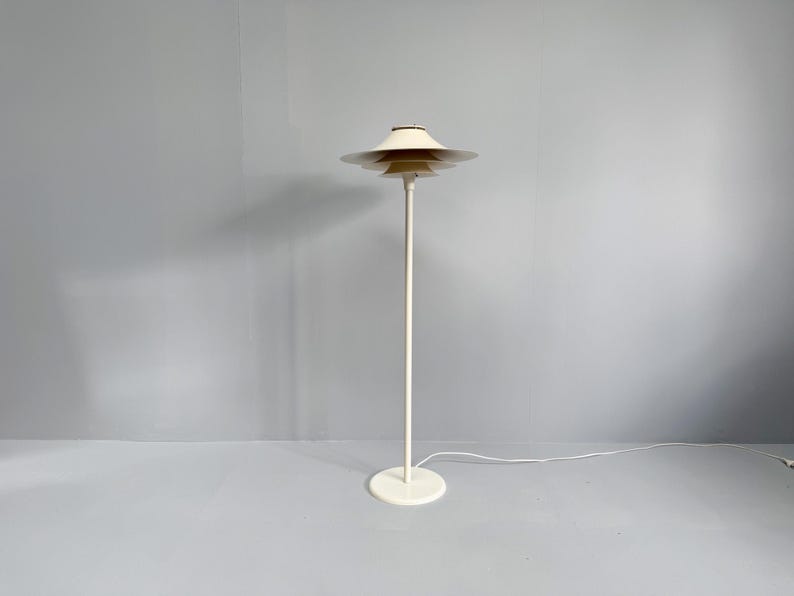 K&ouml;nnte beinhalten: Eine cremefarbene Stehlampe mit einem mehrstufigen Schirmdesign. Die Lampe hat einen schlanken, vertikalen Pfosten und einen kreisf&ouml;rmigen Sockel. Der Schirm besteht aus mehreren gestapelten, scheibenf&ouml;rmigen Elementen. Die Lampe ist an eine Steckdose angeschlossen.