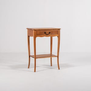 May include: A small, antique wooden side table with a single drawer and a lower shelf. The table has curved legs and a dark handle on the drawer. The wood has a warm, natural finish, and the table is set against a white background.