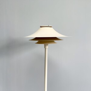 K&ouml;nnte beinhalten: Eine cremefarbene Stehlampe mit einem mehrschichtigen Schirmdesign. Die Lampe hat einen schlanken, cremefarbenen Pfosten und drei gestapelte, tellerf&ouml;rmige Schirme. Die Lampe ist ein modernes Design, und die Gesamt&auml;sthetik ist minimalistisch und elegant.