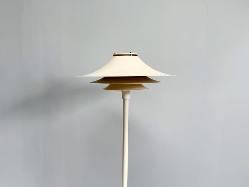 K&ouml;nnte beinhalten: Eine cremefarbene Stehlampe mit einem mehrschichtigen Schirmdesign. Die Lampe hat einen schlanken, cremefarbenen Pfosten und drei gestapelte, tellerf&ouml;rmige Schirme. Die Lampe ist ein modernes Design, und die Gesamt&auml;sthetik ist minimalistisch und elegant.