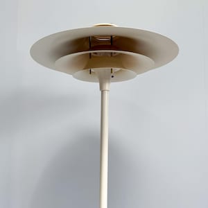 K&ouml;nnte beinhalten: Eine wei&szlig;e Stehlampe mit einem mehrstufigen Schirmdesign. Die Lampe hat einen hohen, schlanken Pfosten und eine Reihe von kreisf&ouml;rmigen Schirmen, die sich nach oben verkleinern. Die Lampe hat eine neutrale Farbe und eine moderne &Auml;sthetik.