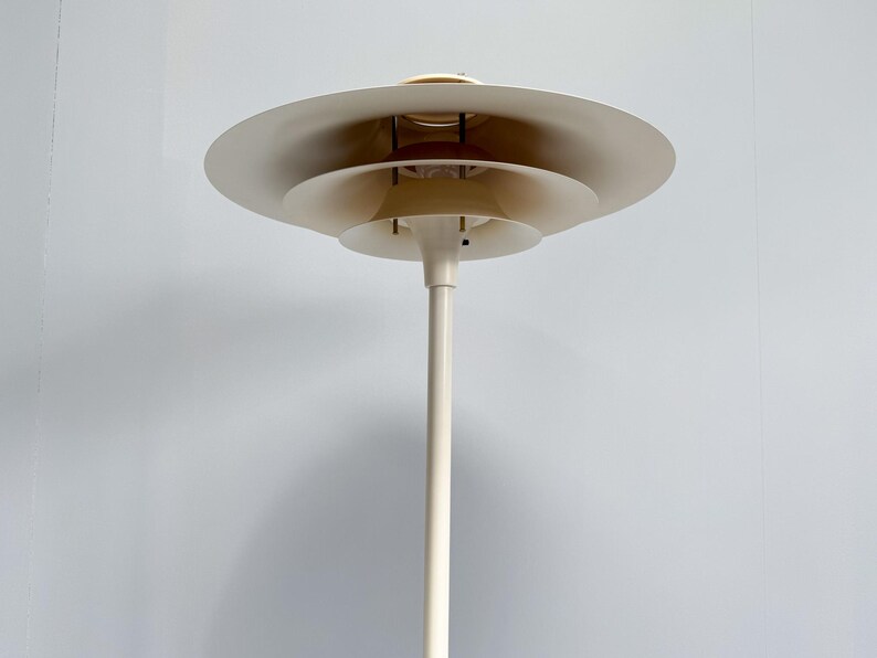K&ouml;nnte beinhalten: Eine wei&szlig;e Stehlampe mit einem mehrstufigen Schirmdesign. Die Lampe hat einen hohen, schlanken Pfosten und eine Reihe von kreisf&ouml;rmigen Schirmen, die sich nach oben verkleinern. Die Lampe hat eine neutrale Farbe und eine moderne &Auml;sthetik.