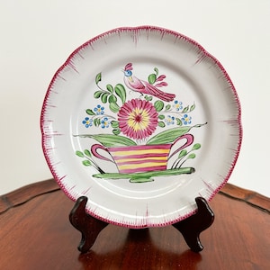 Könnte beinhalten: Ein weißer Keramikteller mit rosafarbenem Rand und einem handgemalten Design eines rosa Vogels, der auf einer Blume in einem gestreiften Topf sitzt. Der Teller steht auf einem Holzständer.