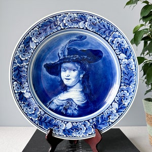 May include: A blue and white Delftware plate with a painted portrait of a woman wearing a hat. The plate is decorated with a floral border.