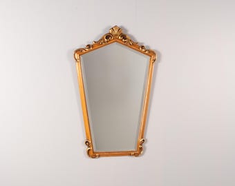 May include: A wall mirror with a gold-toned frame featuring a tapered, pentagonal shape. The top of the frame is adorned with a decorative shell-like motif and scrollwork, while the bottom corners have subtle, rounded scroll accents.