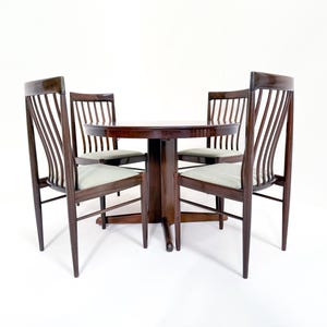 Può includere: Un set da pranzo in legno scuro con un tavolo rotondo e quattro sedie. Le sedie hanno cuscini di seduta verde chiaro e uno schienale a doghe. Il set è su sfondo bianco.
