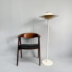 K&ouml;nnte beinhalten: Innenaufnahme mit einem Holzstuhl mit schwarzem Sitz und einer wei&szlig;en Stehlampe. Die Lampe hat einen mehrschichtigen Schirm und einen runden Sockel. Der Stuhl hat eine geschwungene R&uuml;ckenlehne und vier Beine. Die Lampe ist ca. 150 cm hoch.