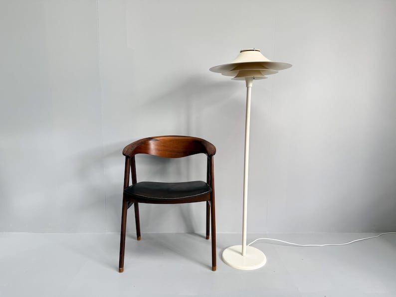 K&ouml;nnte beinhalten: Innenaufnahme mit einem Holzstuhl mit schwarzem Sitz und einer wei&szlig;en Stehlampe. Die Lampe hat einen mehrschichtigen Schirm und einen runden Sockel. Der Stuhl hat eine geschwungene R&uuml;ckenlehne und vier Beine. Die Lampe ist ca. 150 cm hoch.