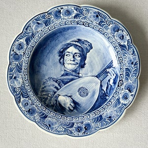 May include: A blue and white ceramic plate with a decorative border of flowers and swirls. The center of the plate features a painting of a person playing a lute.