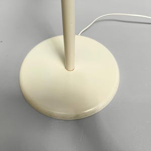 K&ouml;nnte beinhalten: Nahaufnahme eines cremefarbenen Stehlampenfu&szlig;es und -stiels. Der kreisf&ouml;rmige Sockel ist massiv und weist ein leicht abgenutztes Aussehen auf. Ein wei&szlig;es Stromkabel erstreckt sich vom Sockel.