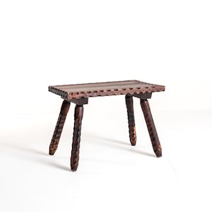 Puede incluir: Un pequeño taburete de madera hecho a mano con una parte superior rectangular y tres patas. El taburete marrón oscuro presenta detalles tallados en las patas y los bordes, mostrando una estética rústica. El taburete mide aproximadamente 30 cm de largo.