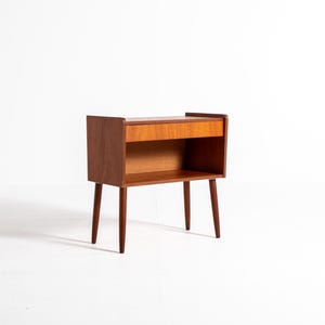 May include: A mid-century modern wooden bedside table with a drawer and open shelf. The table is made of warm-toned wood and has tapered legs. The dimensions are approximately 61 cm high, 51 cm wide, and 36 cm deep.