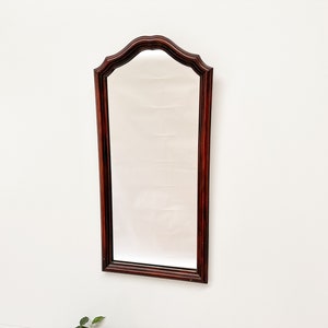 Puede incluir: Un espejo alto y rectangular con un marco de madera oscura. El marco tiene una parte superior curva y un diseño ligeramente ornamentado.