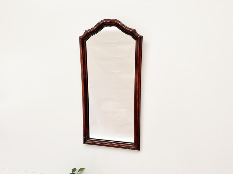 May include: A tall, rectangular mirror with a dark wood frame. The frame has a curved top and a slightly ornate design.