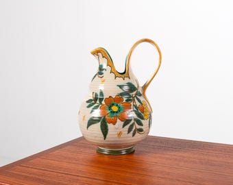 Puede incluir: Una jarra de cerámica con base crema, adornada con diseños florales naranjas, amarillos y verdes. La jarra tiene un asa curva y un pico con ribete dorado. La jarra mide aproximadamente 25 cm de altura.