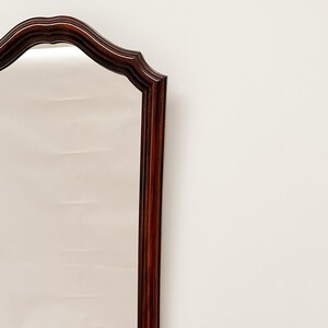 May include: A tall, rectangular mirror with a dark wood frame. The frame has a decorative, arched top with a scalloped edge.