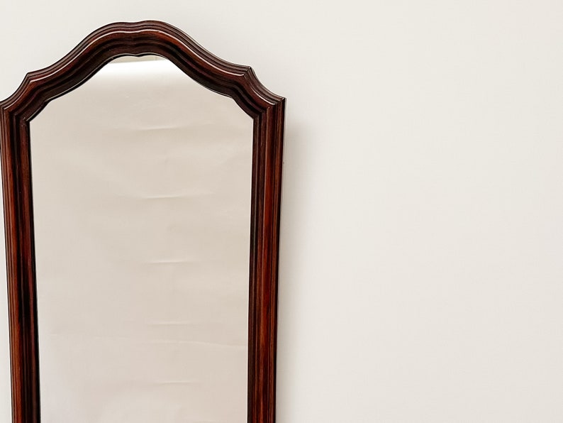May include: A tall, rectangular mirror with a dark wood frame. The frame has a decorative, arched top with a scalloped edge.