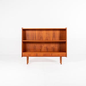 May include: A mid-century modern wooden bookcase with three drawers and two shelves. The bookcase is made of a warm-toned wood and stands on four legs. The design is simple and functional, perfect for storage and display.