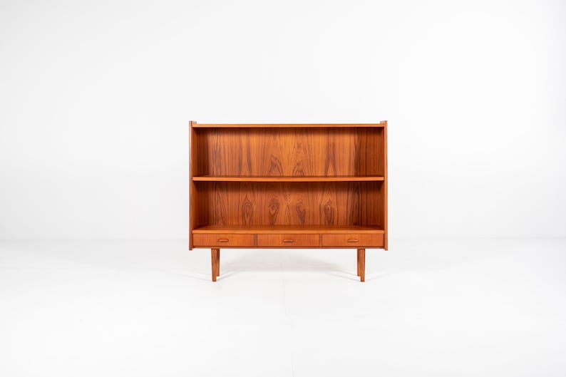 May include: A mid-century modern wooden bookcase with three drawers and two shelves. The bookcase is made of a warm-toned wood and stands on four legs. The design is simple and functional, perfect for storage and display.
