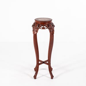 May include: A tall, dark reddish-brown wooden pedestal stand featuring a circular top with an inlaid stone surface. The stand has four curved legs connected by a cross-shaped stretcher, with intricate floral carvings decorating the upper apron.